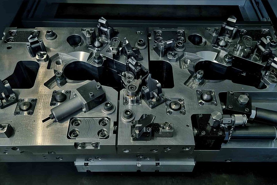 Graue Metallform mit Aussparungen und Befestigungselementen auf dunklem Hintergrund.